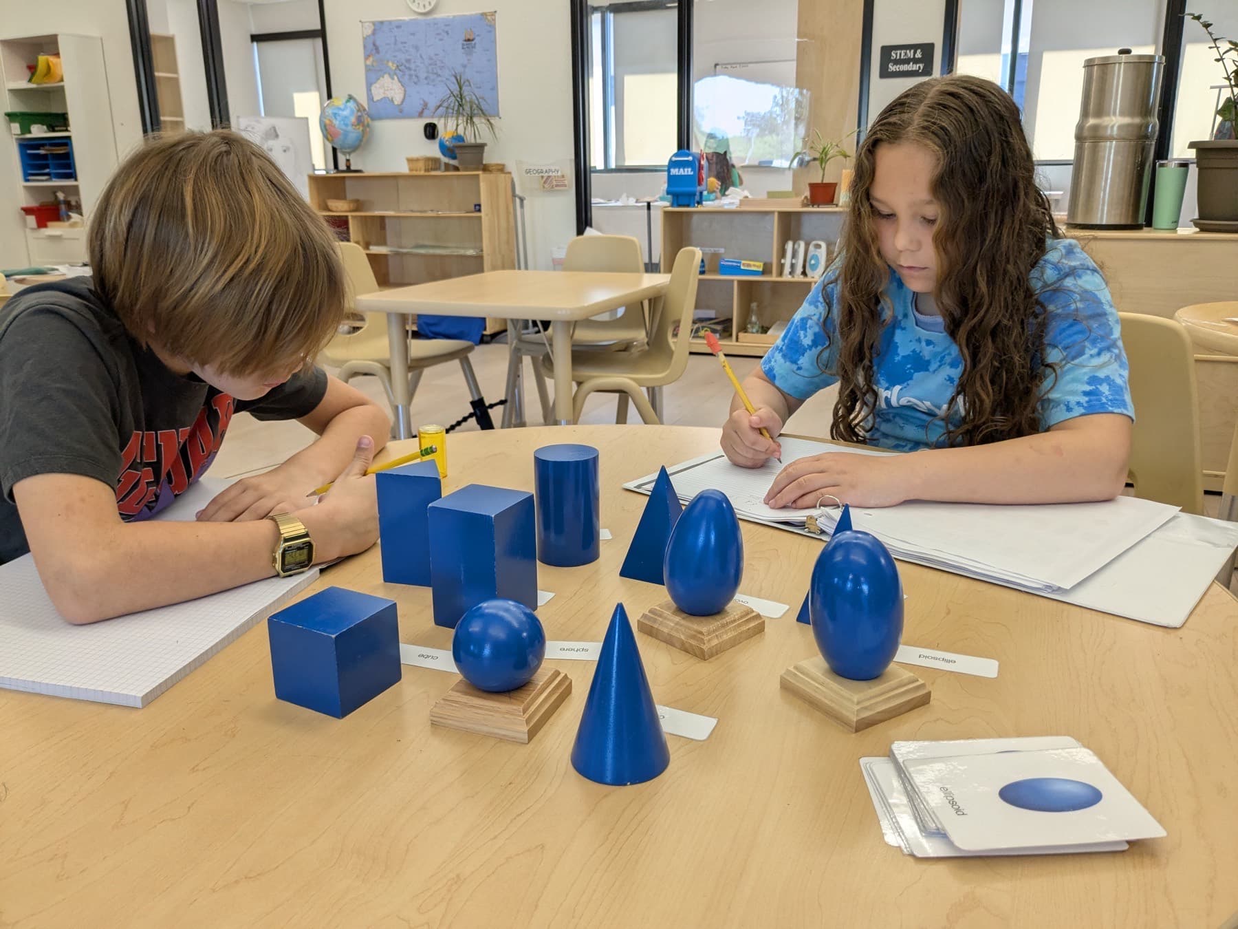 Two students working at a table with wooden geometry solids and lined notebooks